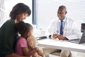 Pediatric patient, with teddy bear on mother's lap. They are facing a doctor. All are underrepresented minorities.
